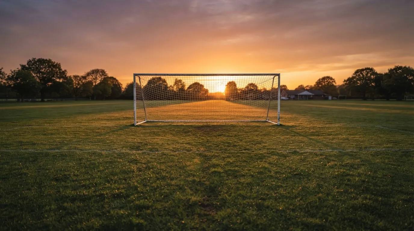 Ein Fußballtor mit Netz bei Sonnenuntergang auf einem leeren Platz, die Schatten der Torpfosten liegen lang auf dem Gras.