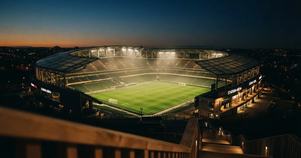 Ein Fußballstadion unter Flutlicht mit hellen Rängen, von einem Hügel aus gesehen, darüber ein dunkelblauer Abendhimmel.