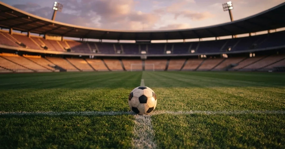 Ein Fußball liegt auf dem Anstoßpunkt eines leeren Rasenplatzes, im Hintergrund eine große Stadiontribüne im Abendlicht.