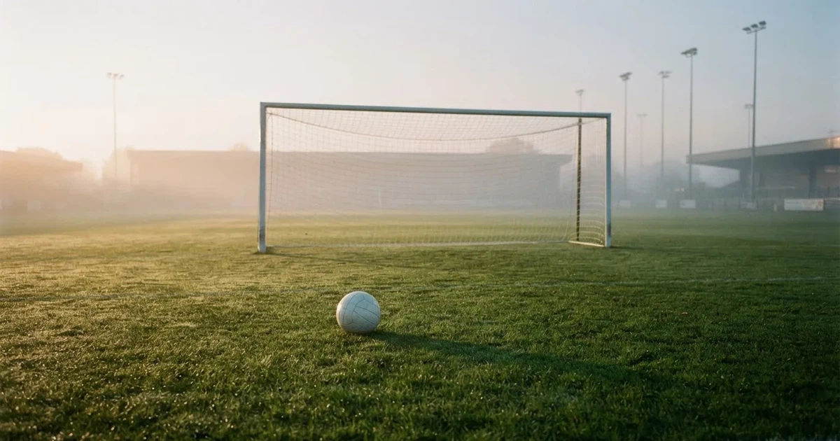 Ein Fußballtor mit gespanntem Netz steht auf einem leeren Rasenplatz, davor ein einzelner Ball, Morgendunst liegt über dem Feld.