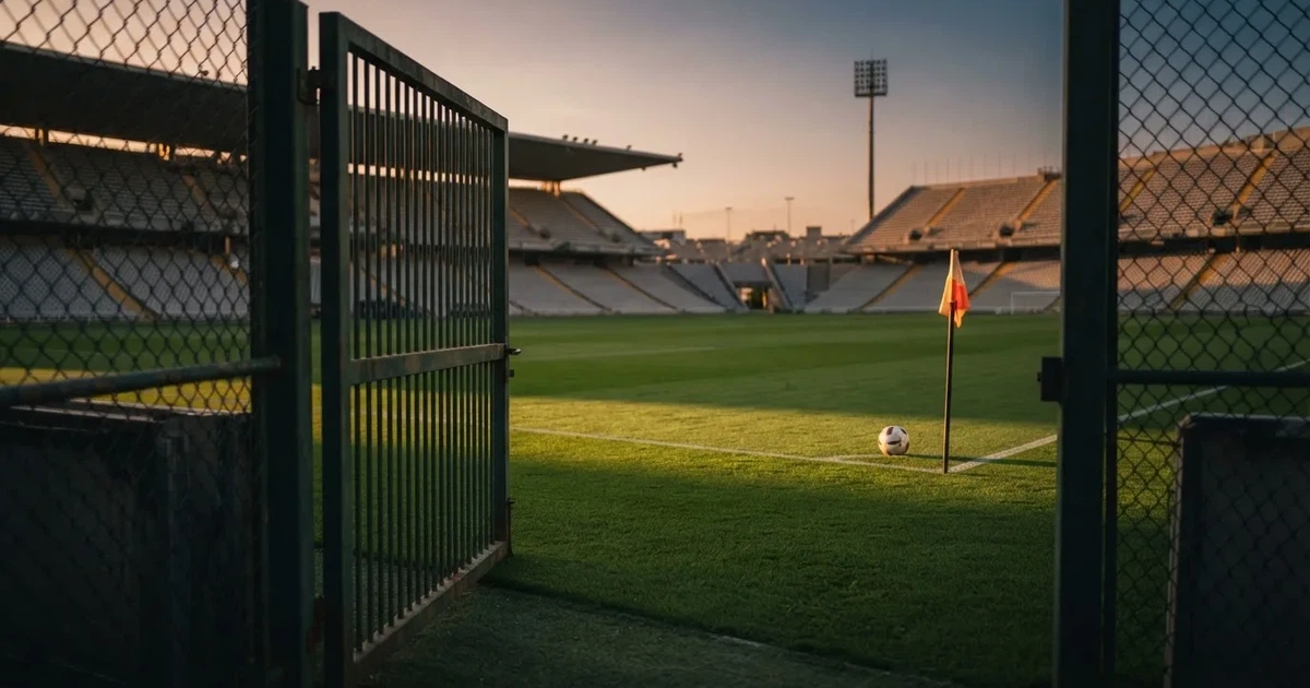 Blick durch ein Stadiontor auf einen grünen Rasen im Abendlicht, ein Ball liegt an der Eckfahne, die Ränge sind leer.