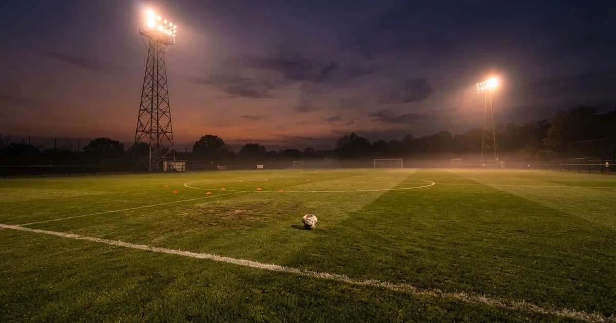 Eine Flutlichtanlage ragt über einen Trainingsplatz, auf dem Rasen liegen verstreut Hütchen und ein Ball, am Himmel die Abenddämmerung.