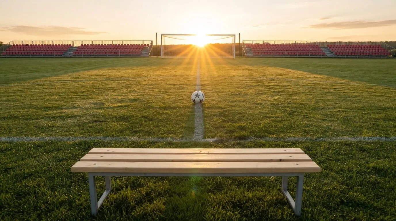 Ein Fußball auf einem Rasenplatz im Gegenlicht der tiefstehenden Sonne, am Rand eine leere Ersatzbank.
