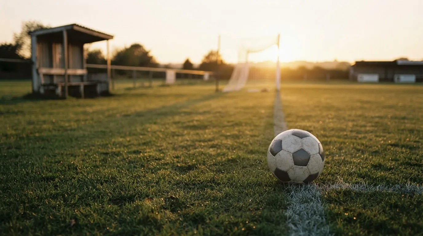 Ein Fußball liegt im Gras am Spielfeldrand, dahinter eine leere Bank und ein Tor im Gegenlicht.