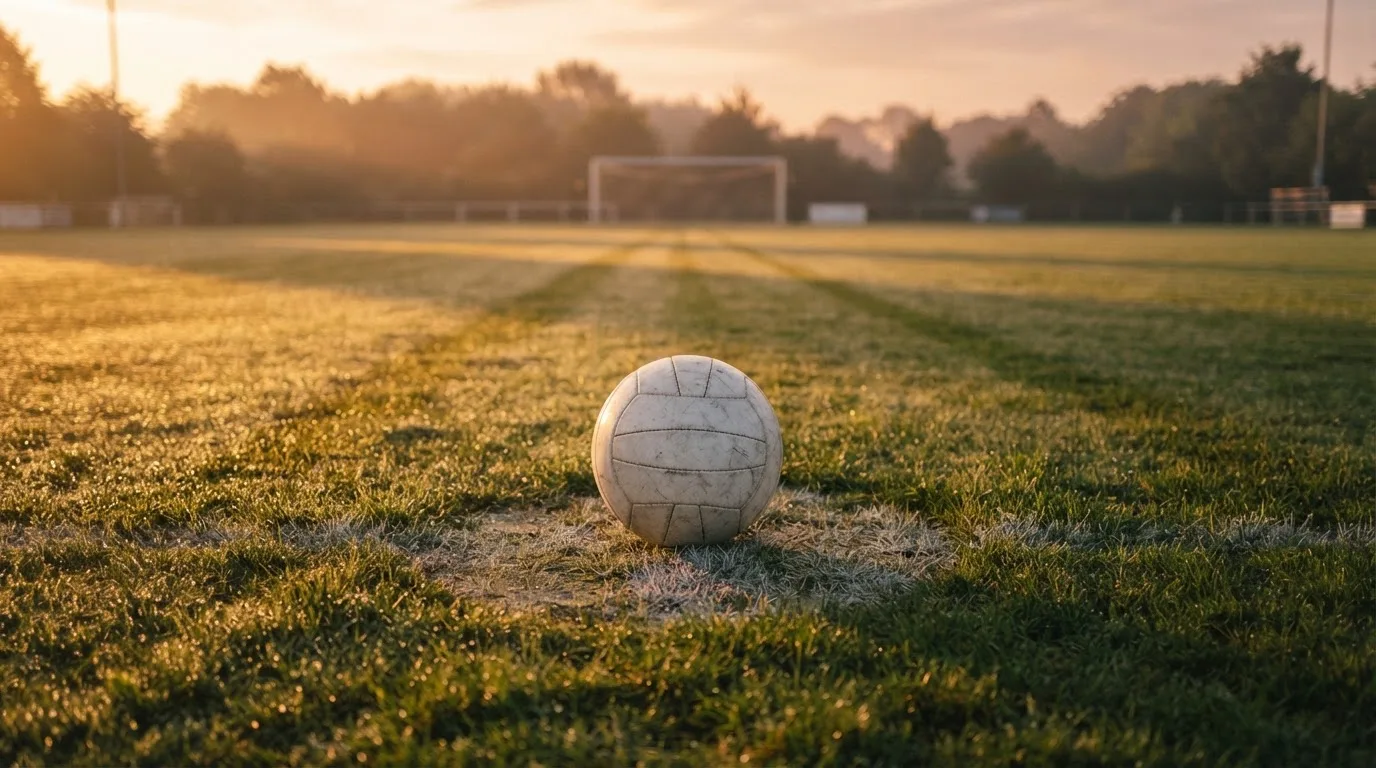 Ein Fußball liegt am Rand des Mittelkreises auf einem leeren Rasenplatz bei Sonnenaufgang.