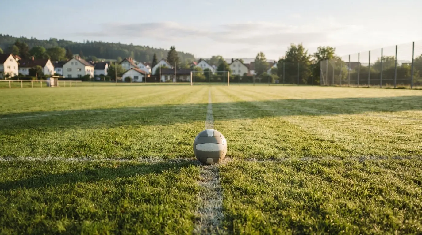 Ein Fußball auf dem Rasen eines leeren Sportplatzes am Rand einer Kleinstadt im Morgenlicht.