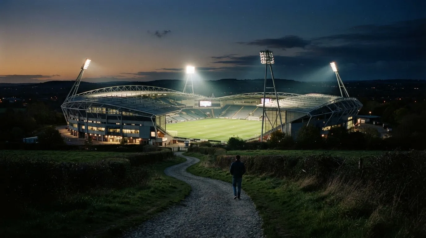 Ein Stadion unter Flutlicht, von einer Anhöhe aus gesehen, davor ein stiller Weg in der Dämmerung.