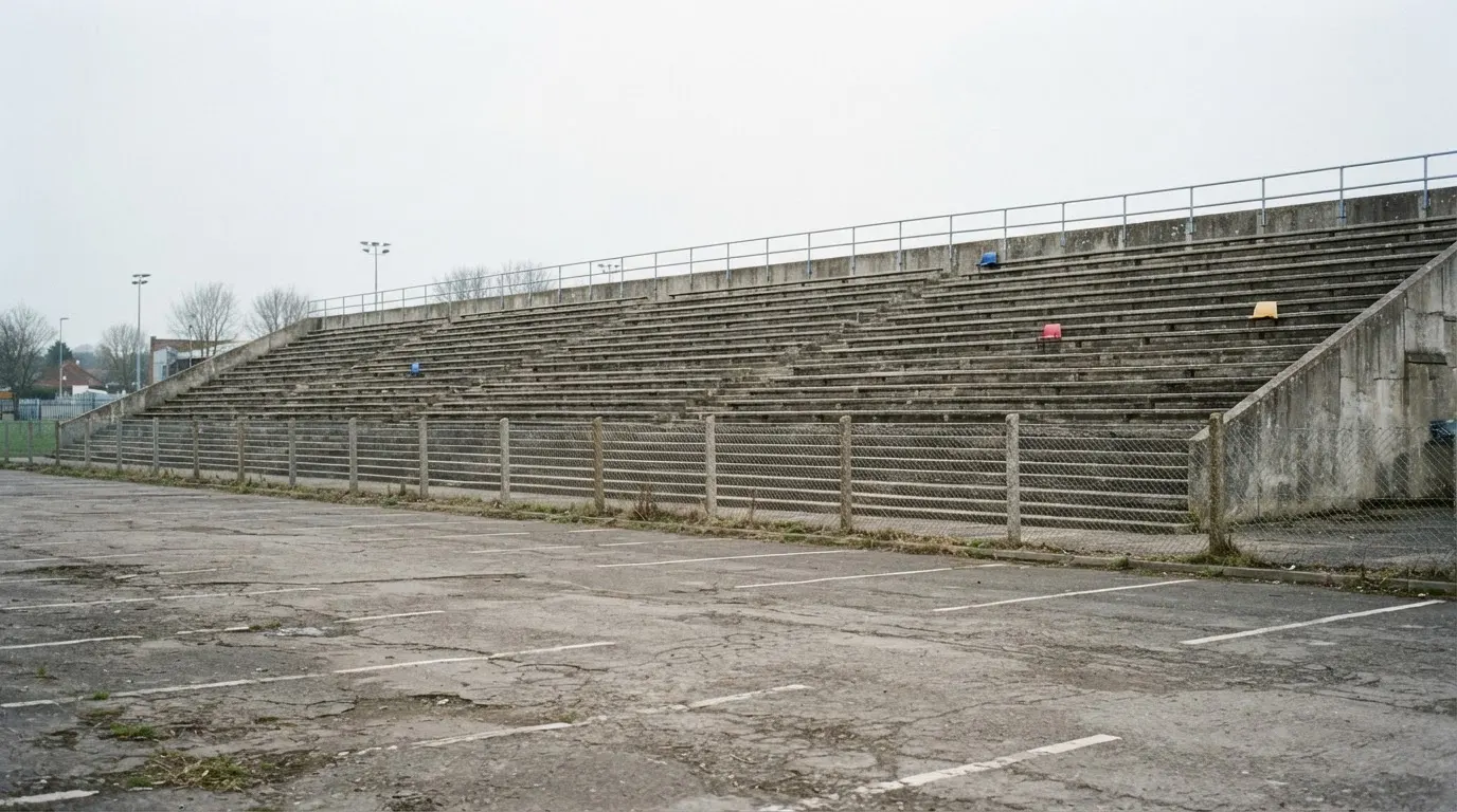 Eine leere Tribüne eines kleinen Stadions am frühen Morgen, aus der Ferne über einen menschenleeren Parkplatz fotografiert.