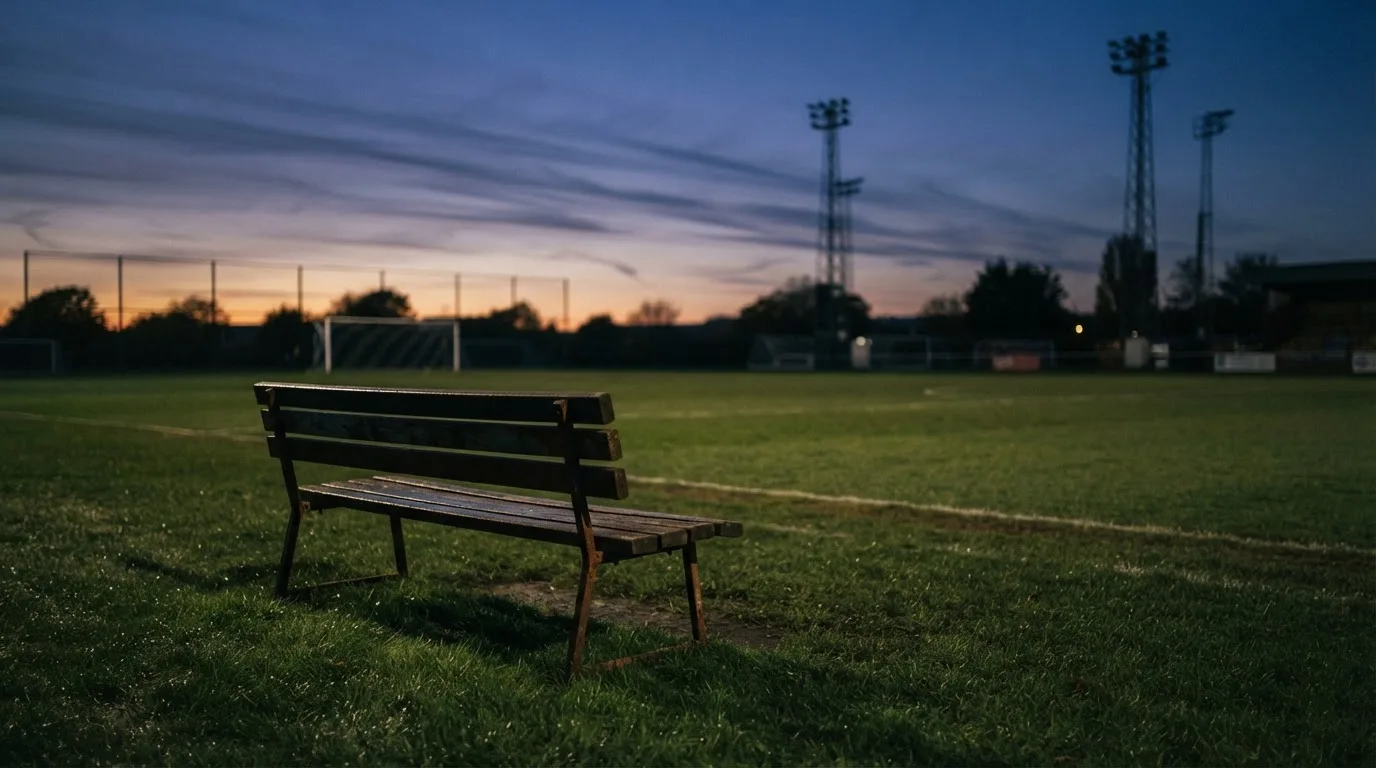 Eine leere Parkbank am Rand eines Fußballfelds in der Abenddämmerung, im Hintergrund ragen dunkle Flutlichtmasten in den Himmel.