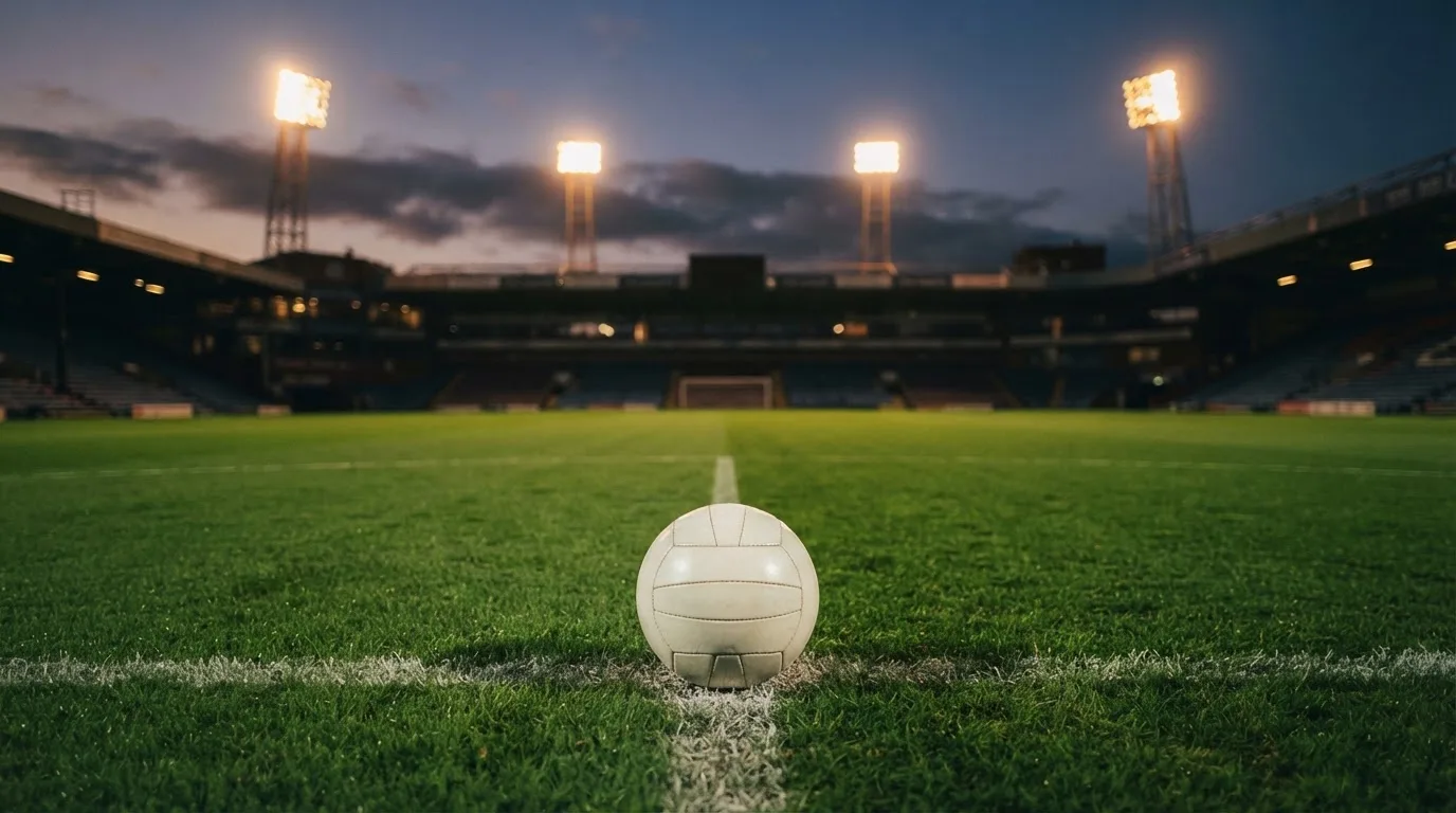Ein Fußball liegt auf dem Anstoßpunkt eines leeren Rasenplatzes, im Hintergrund steht ein Stadion unter Flutlicht in der Dämmerung.