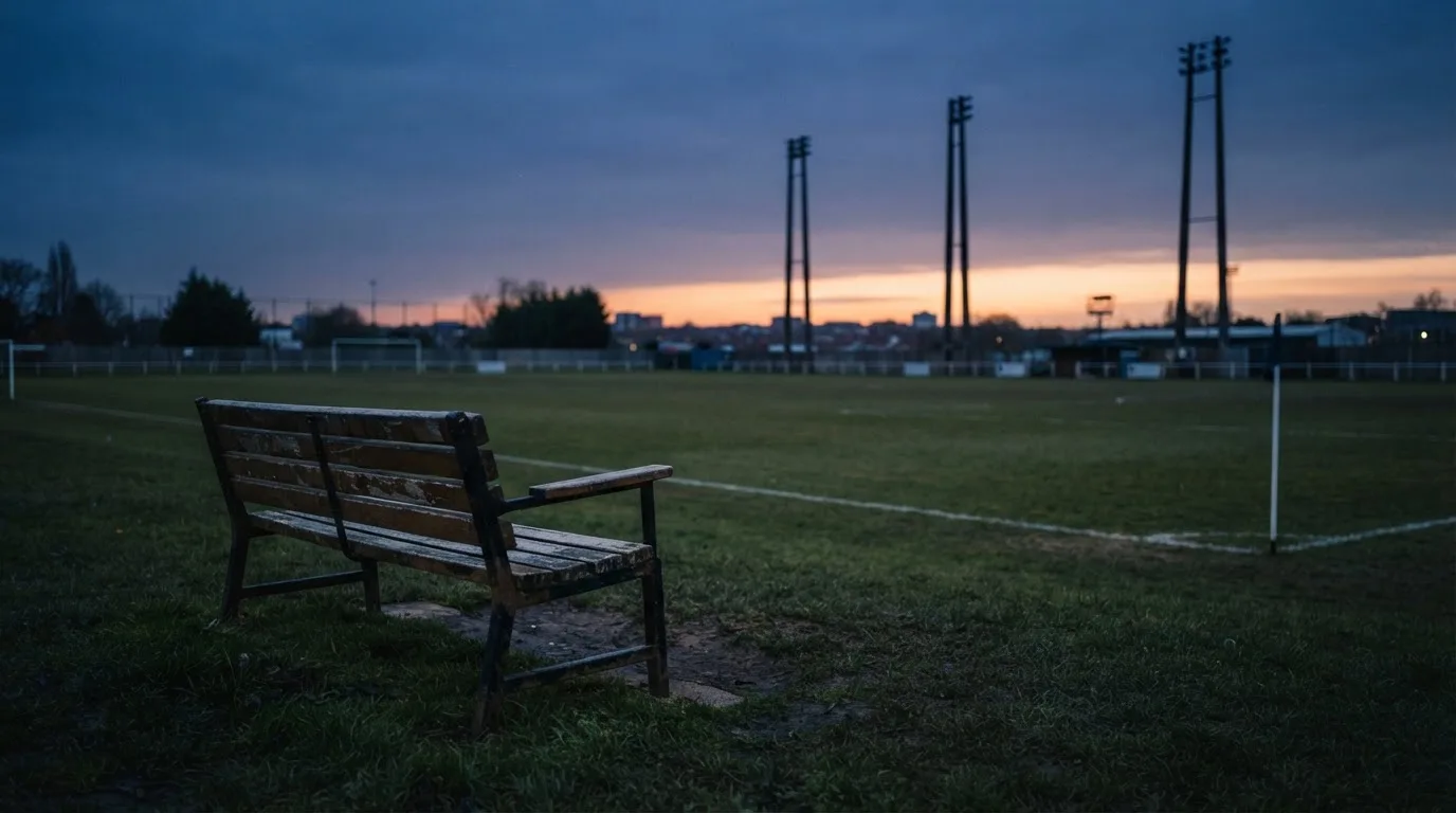 Eine leere Parkbank am Rand eines Fußballfelds in der Abenddämmerung, im Hintergrund ragen dunkle Flutlichtmasten in den Himmel.