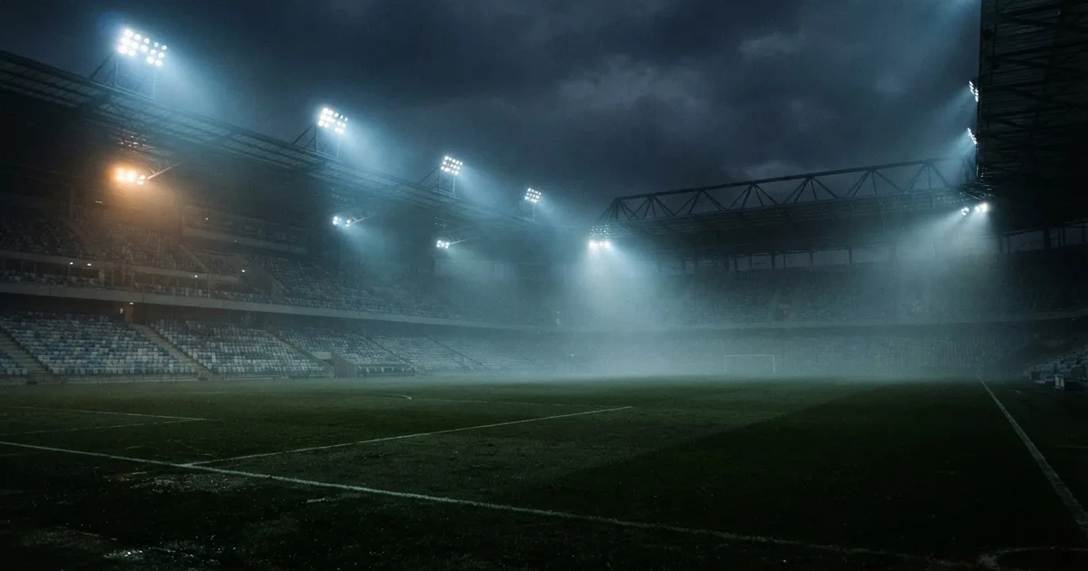 Ein Stadion unter Flutlicht, aus großer Entfernung über eine dunkle Wiese fotografiert, am Himmel zieht Nebel auf.
