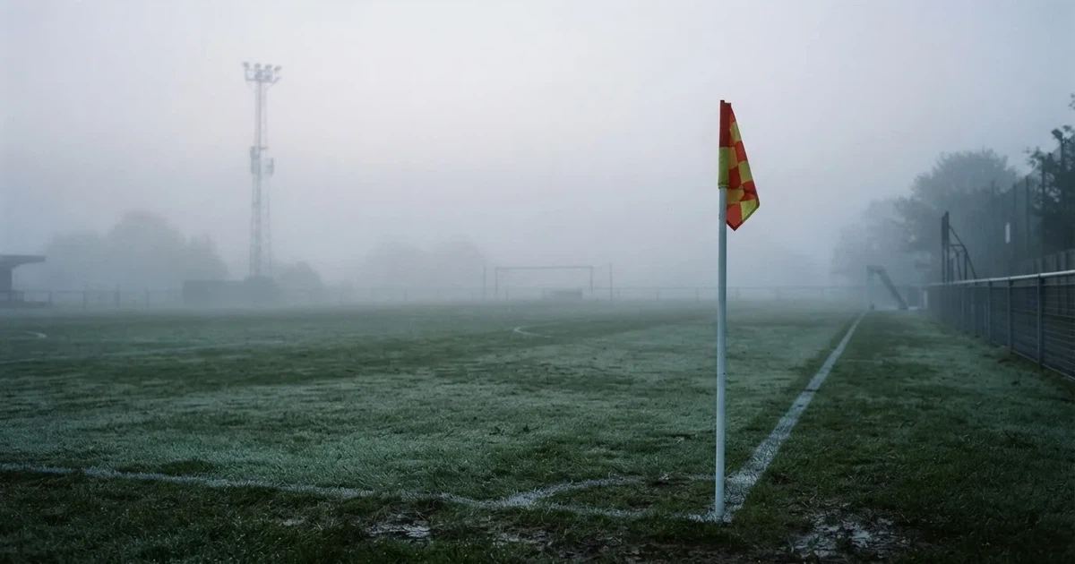 Eine Eckfahne steht im Nebel am Rand eines leeren Rasenplatzes in der Morgendämmerung, dahinter ein Flutlichtmast.