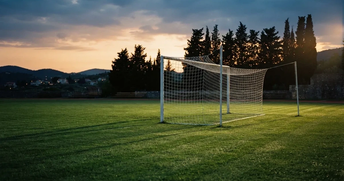 Ein Fußballtor mit Netz auf einem leeren Platz im Abendlicht, der Rasen frisch gemäht, im Hintergrund Zypressen.