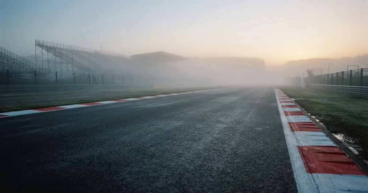 Eine leere Rennstrecke im Morgennebel, Asphalt mit rot-weißen Randsteinen, im Hintergrund Tribünen in der Ferne.