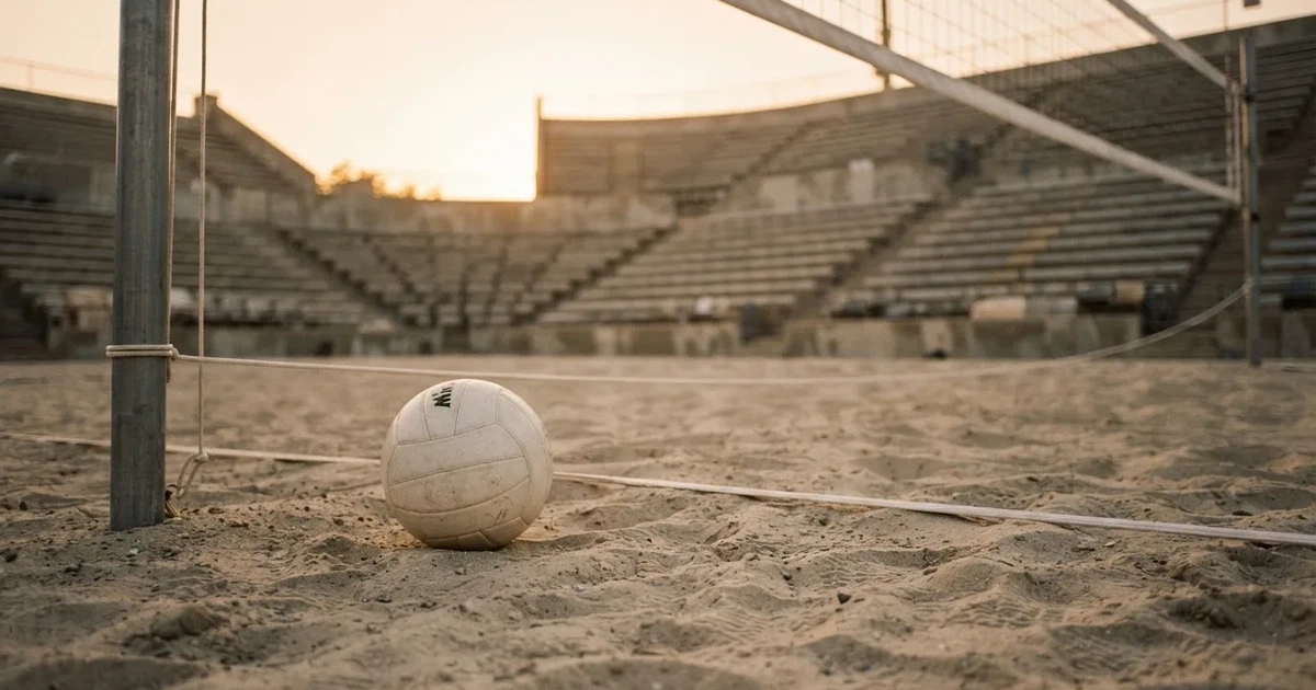Ein Volleyball liegt auf dem Sand eines Beachvolleyball-Feldes neben dem gespannten Netz, im Hintergrund eine leere Tribüne bei Abendlicht.