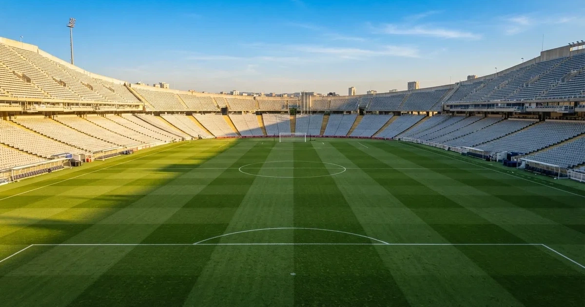 Ein Fußballstadion mit hellen Rängen unter strahlend blauem Himmel, davor ein sonnenbeschienener Rasen mit frisch gemähten Streifen.