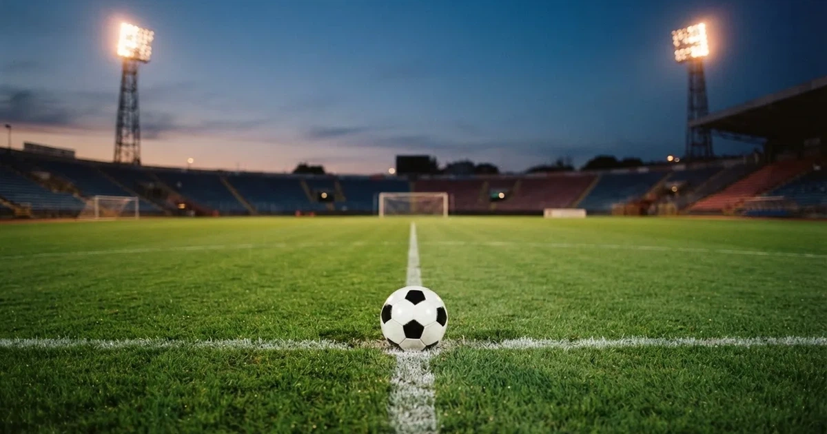 Ein Fußball liegt auf dem Anstoßpunkt eines leeren Rasenplatzes, im Hintergrund eine Tribüne und Flutlichtmasten in der Abenddämmerung.