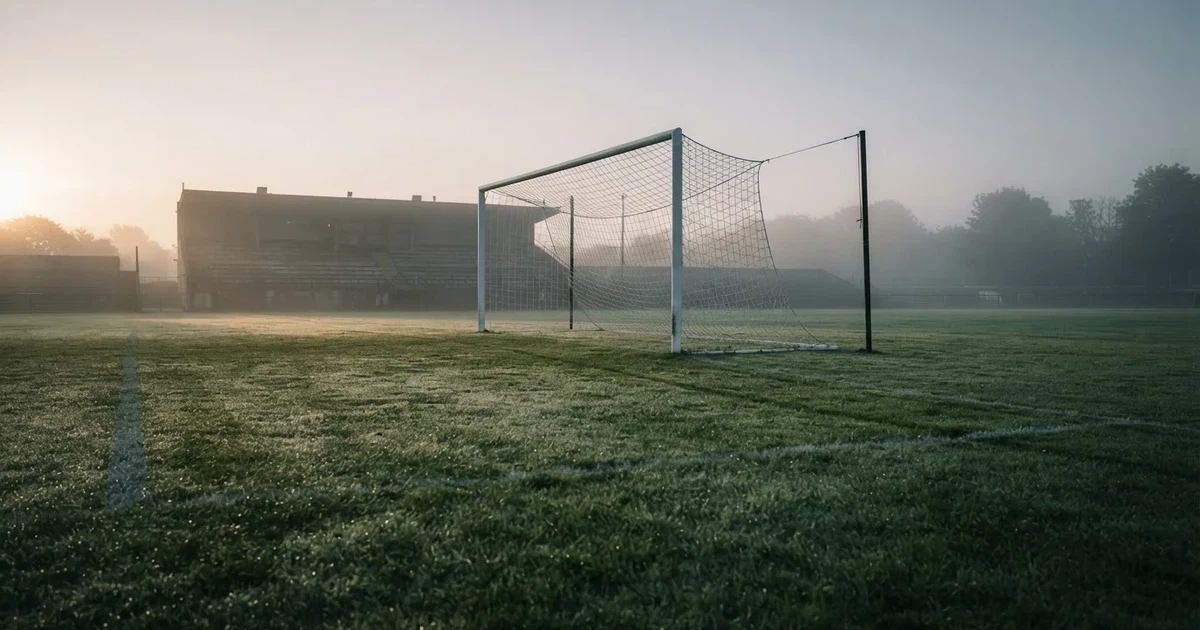 Ein Fußballtor mit Netz steht im Morgennebel auf einem leeren Rasenplatz, im Hintergrund die Silhouette einer kleinen Tribüne.