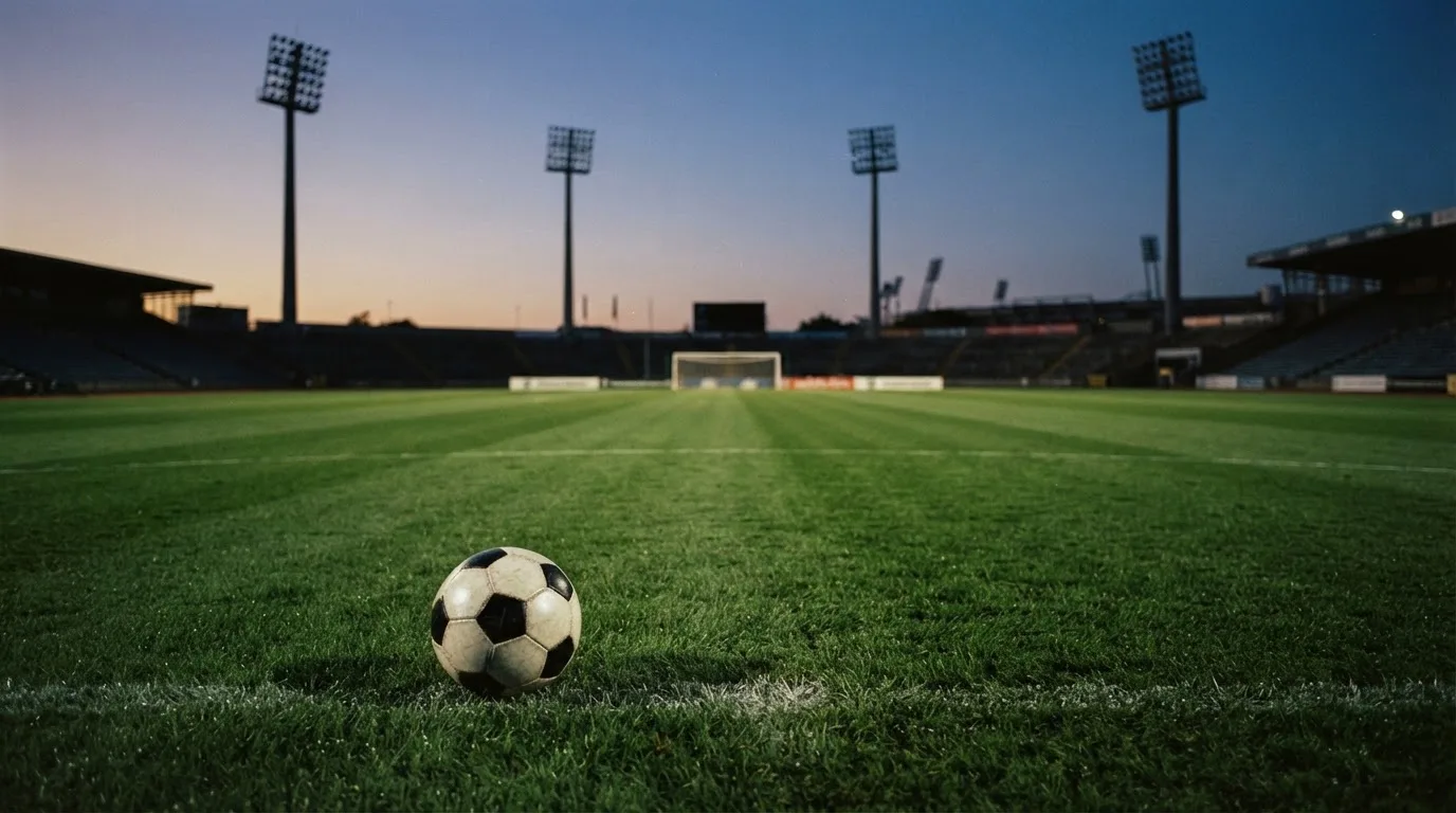 Ein Fußball liegt im Gras eines leeren Platzes, im Hintergrund stehen Flutlichtmasten in der Dämmerung.