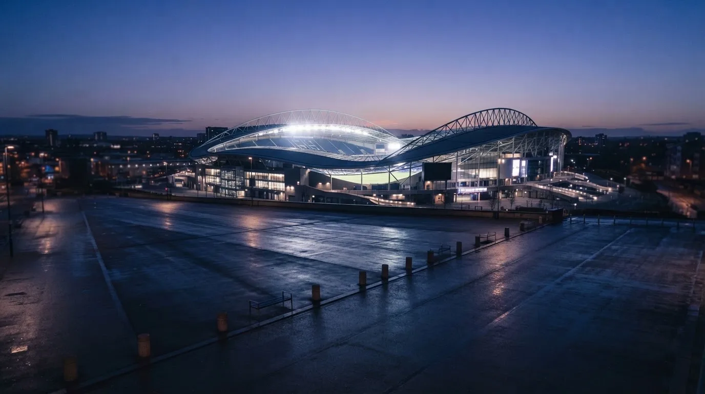 Ein Stadion unter Flutlicht von weitem betrachtet, davor ein stiller Platz bei Dunkelheit und feuchtem Asphalt.