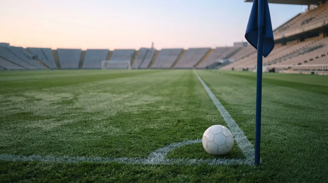 Ein Fußball liegt auf Rasen neben einer Eckfahne ohne Aufdruck auf einem leeren Platz im Morgenlicht.