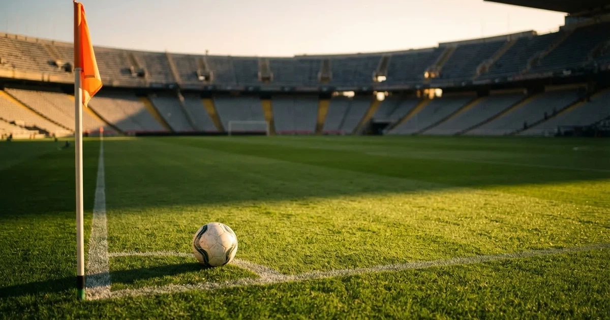 Ein Fußball liegt am Viertelkreis an der Eckfahne eines Rasenplatzes, im Hintergrund leere Tribünen in warmem Abendlicht.