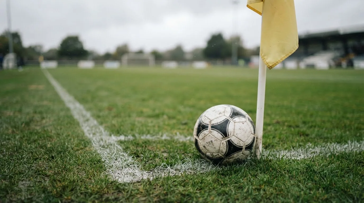 Ein Fußball liegt neben einer Eckfahne auf kurz gemähtem Rasen unter bewölktem Himmel.
